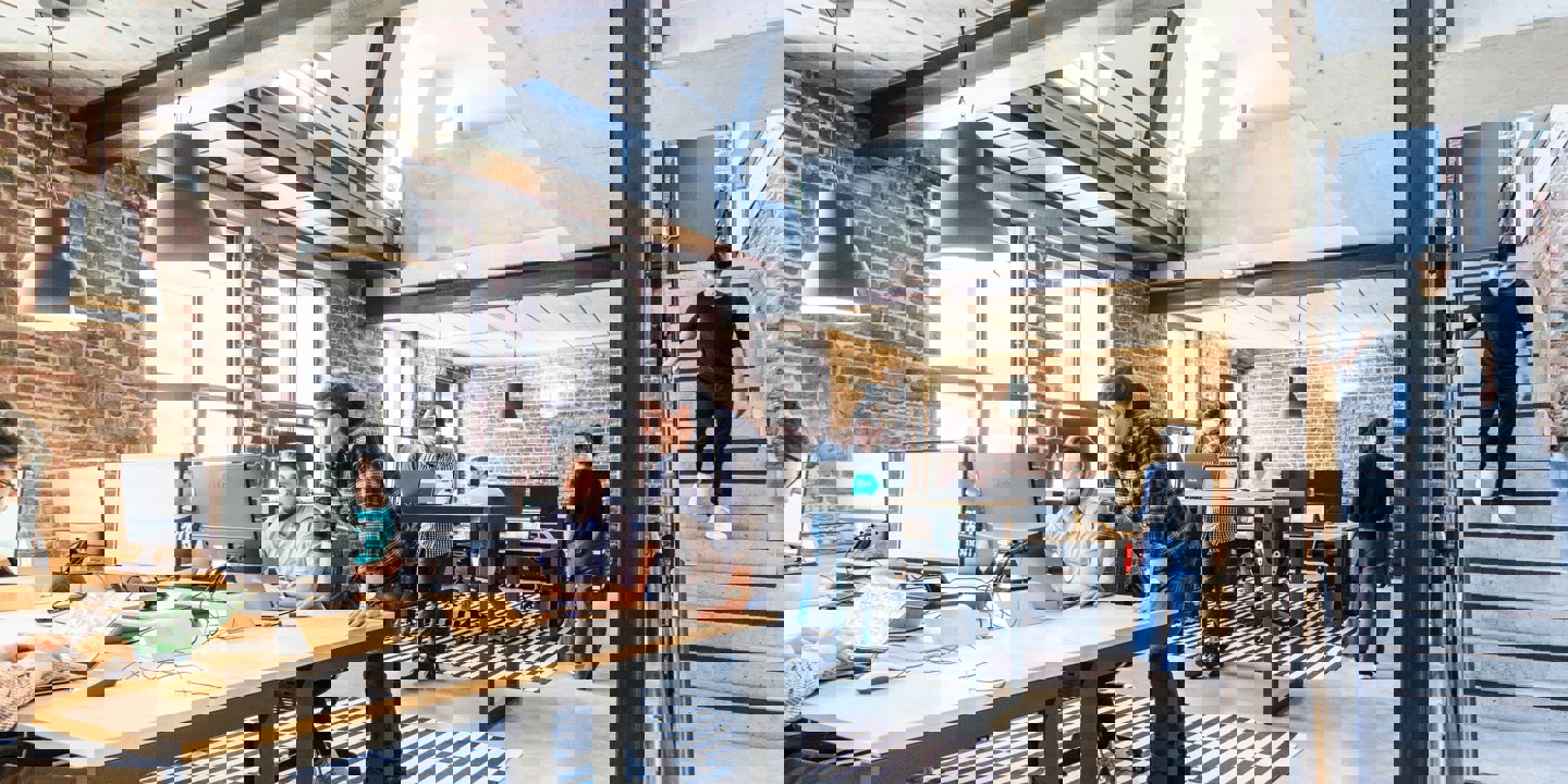 Open plan office with long tables and laptops with brick interior and wooden floor and stairs